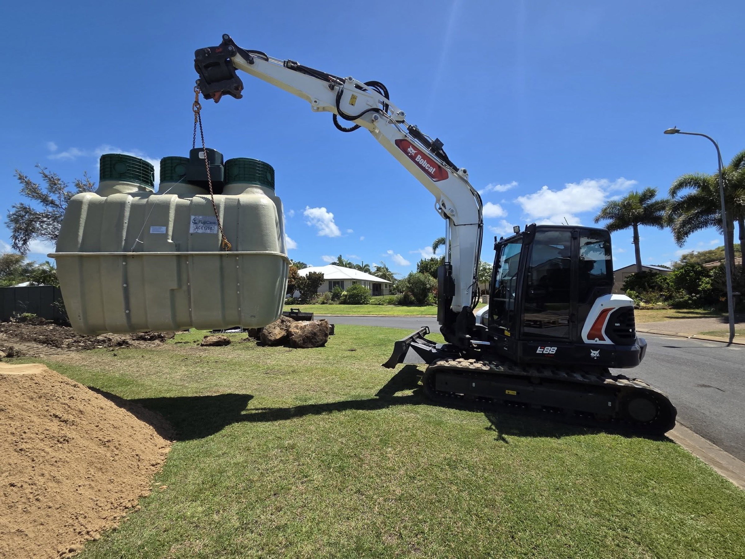 An excavator lifting a wastewater treatment tank beside a suburban street