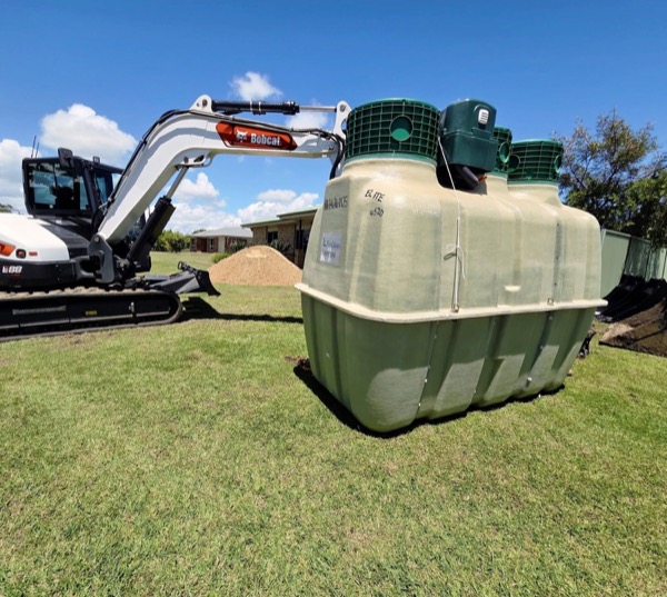An excavator beside a large treatment tank on a grassed block