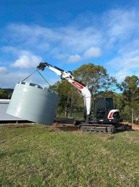 An excavator lifting a round treatment tank across a grassed yard