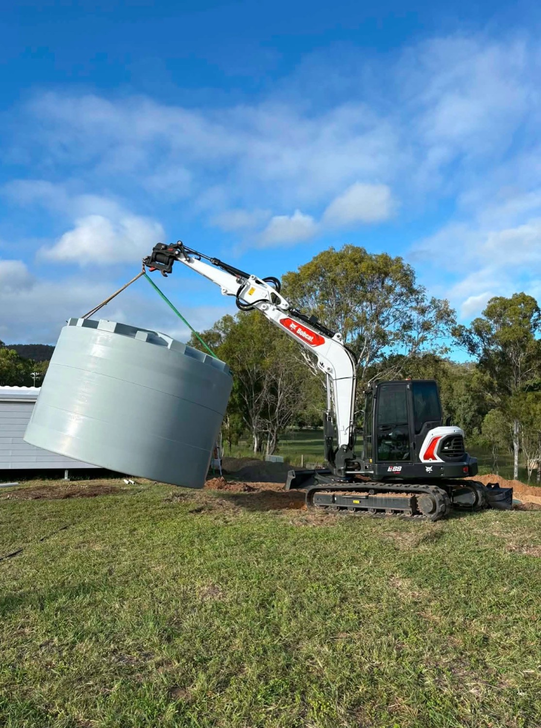 An excavator lifting a round treatment tank across a grassed yard
